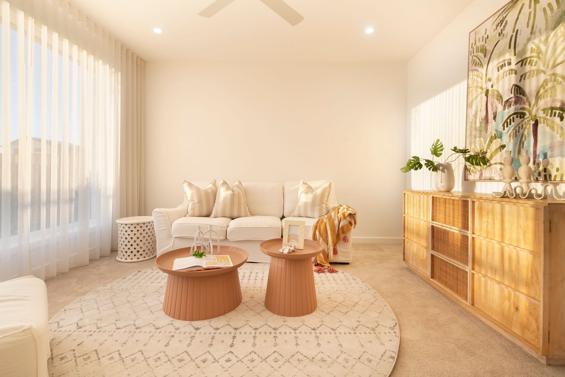 A carpeted sitting room with sheer curtains to two walls, a pale sofa, two round coffee tables and a rattan-fronted timber sideboard.