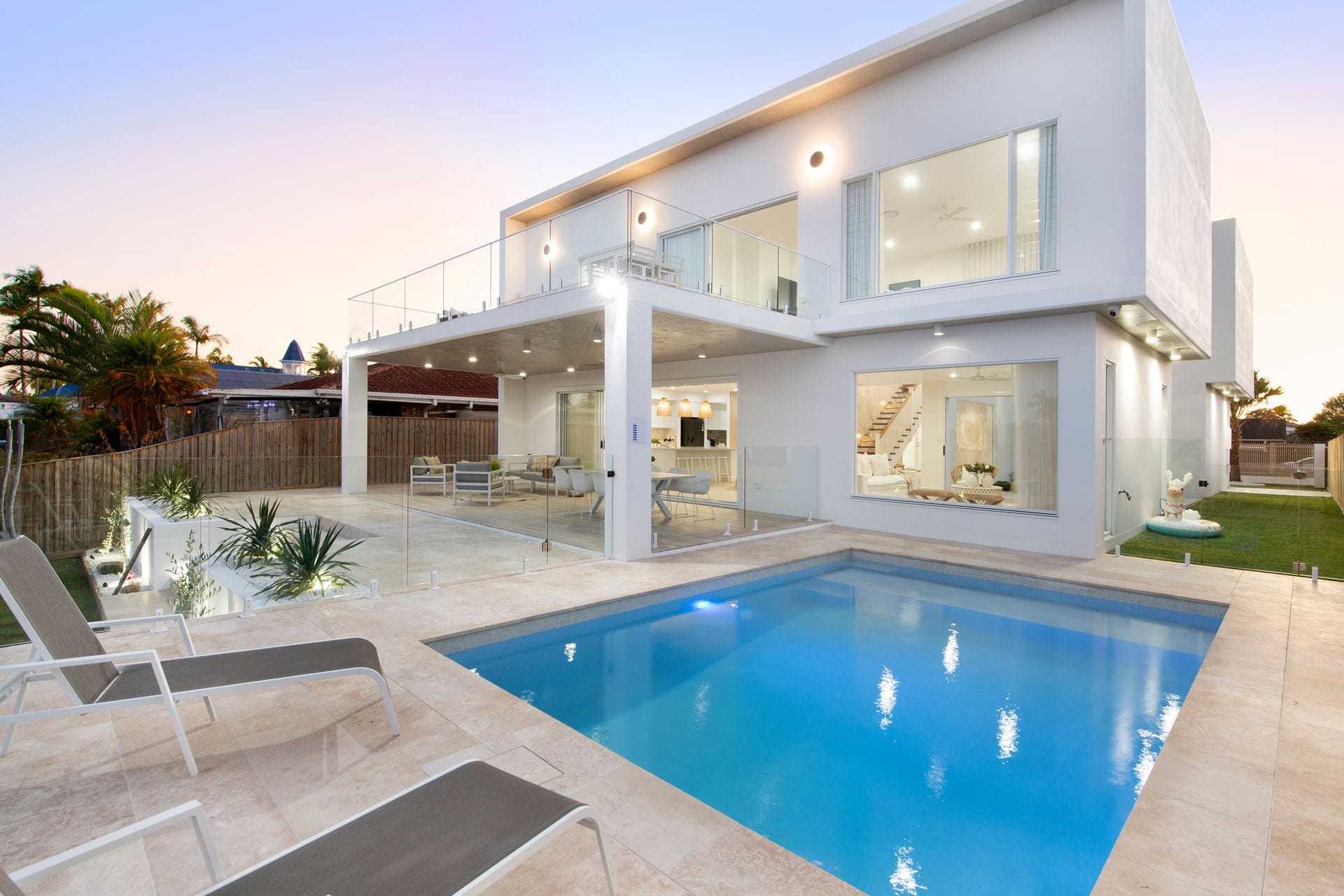 The pool and travertine terrace at dusk, looking back towards the covered alfresco, the full-height glazing behind it and the balcony above.